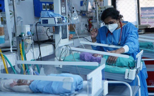 Bildet viser to spedbarn som ligg i små sjukehussenger på eit sjukehus. Ein lege med munnbind og blå sjukehusklede har handa på det eine spedbarnet. Rommet har mykje medisinske apparat, utstyr og ledningar.