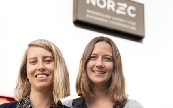 Biletet viser to unge kvinner - ei med lyst hår, ei med mørkt hår. Biletet er teke frå skuldrane og opp. Dei står foran eit kontorbygg, og i bakgrunnen skimtar du Norec-logoen.
